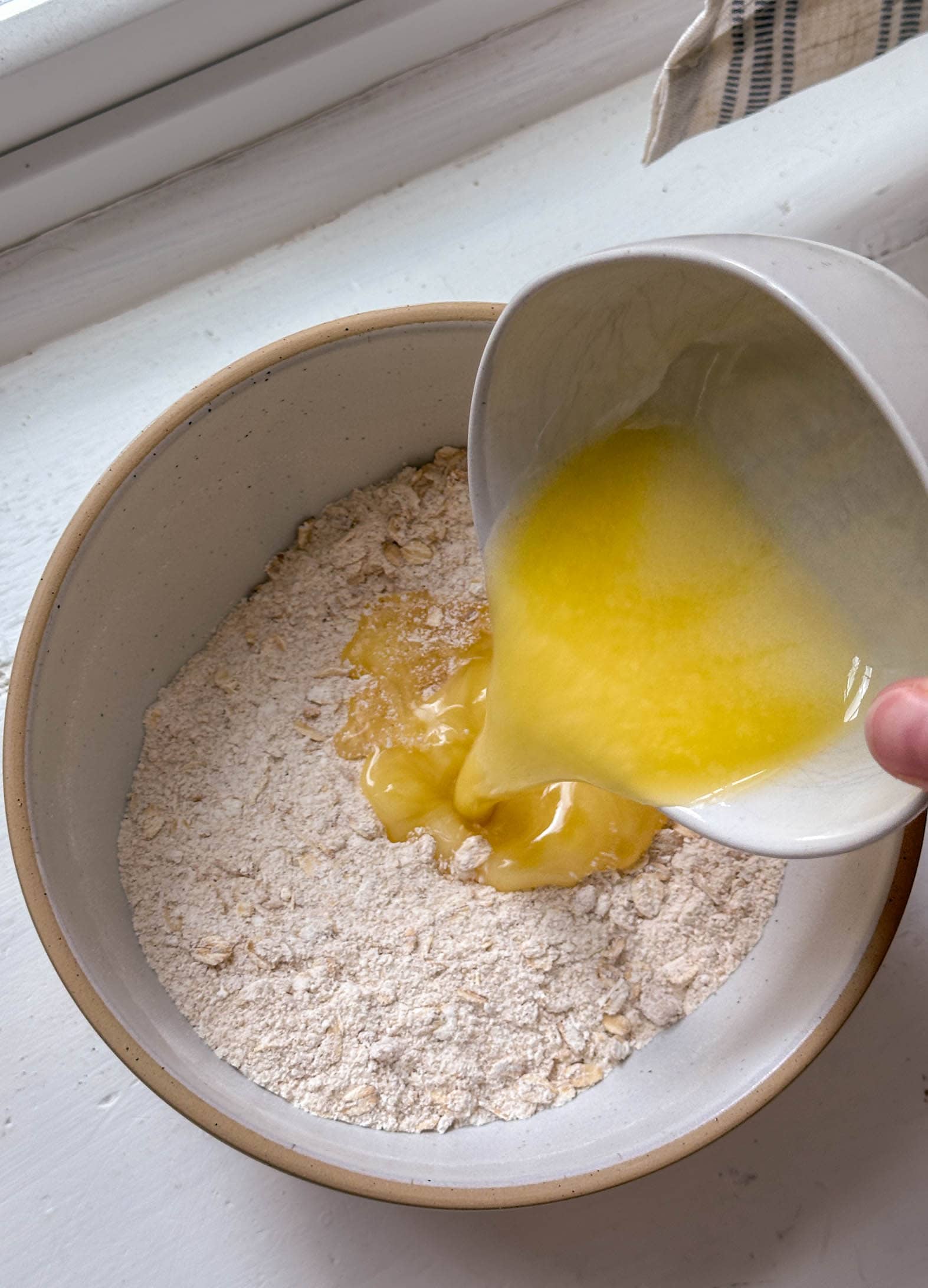 Melted butter is being poured from a white bowl into a bowl of dry ingredients.