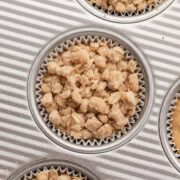 Muffin tin with paper liners, each filled with unbaked crumbly oat topping for muffins or crisps.