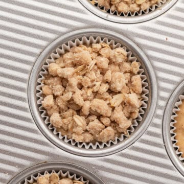 Muffin tin with paper liners, each filled with unbaked crumbly oat topping for muffins or crisps.