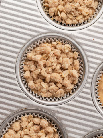 Muffin tin with paper liners, each filled with unbaked crumbly oat topping for muffins or crisps.
