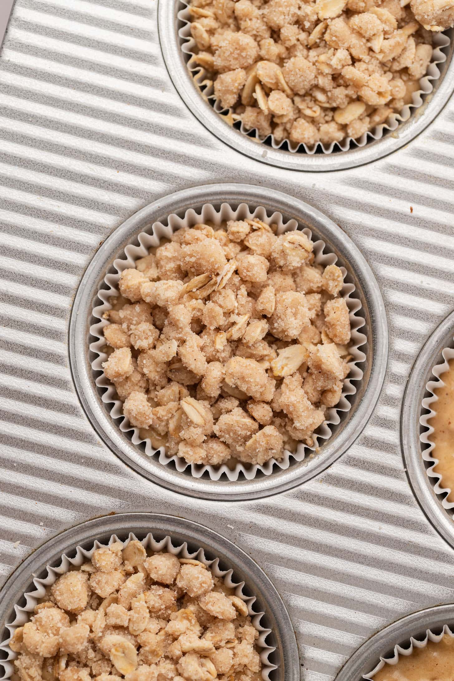 Muffin tin with paper liners, each filled with unbaked crumbly oat topping for muffins or crisps.
