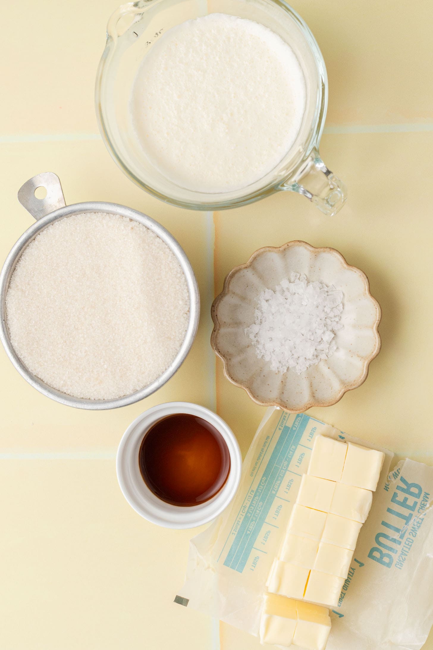 Butter, sugar, vanilla, salt, and cream measured in bowls and cups on a light yellow background.