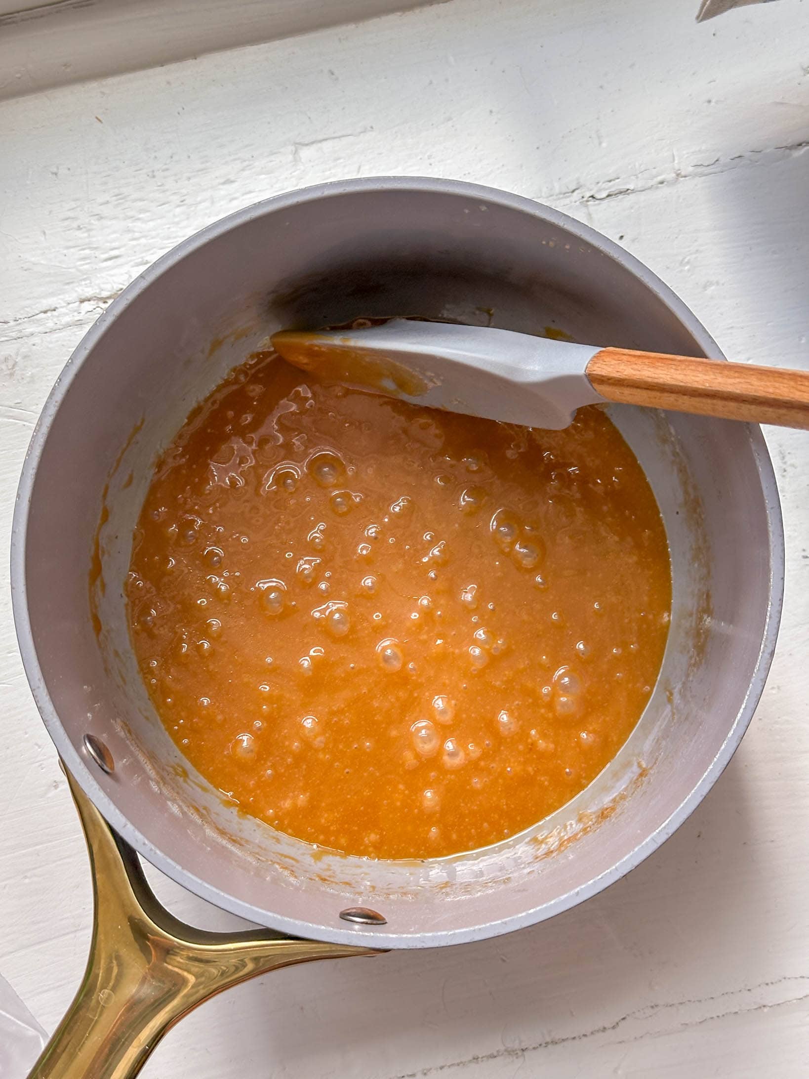 A saucepan of bubbling caramel being stirred with a silicone spatula on a white countertop.