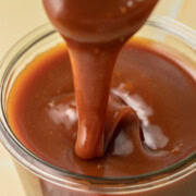 A spoon drizzling thick caramel sauce into a glass jar filled with caramel sauce.