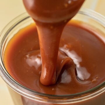 A spoon drizzling thick caramel sauce into a glass jar filled with caramel sauce.