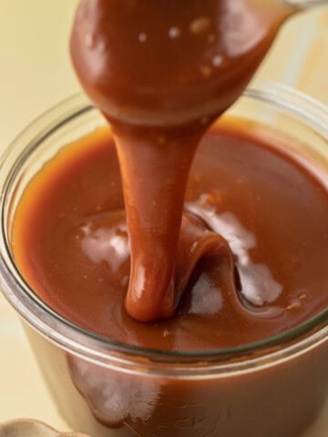 A spoon drizzling thick caramel sauce into a glass jar filled with caramel sauce.