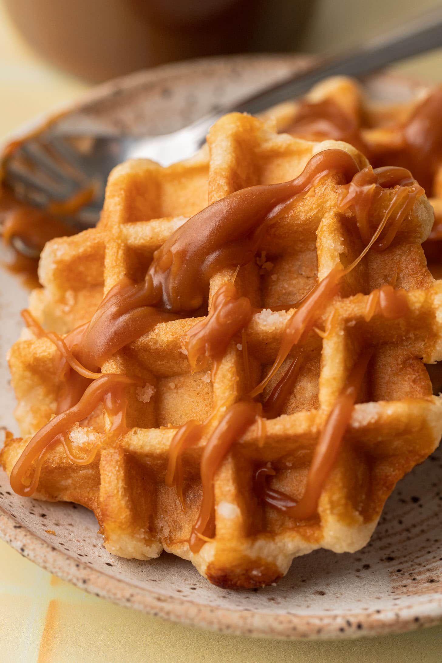 A close-up of a waffle drizzled with caramel sauce, served on a ceramic plate with a fork.