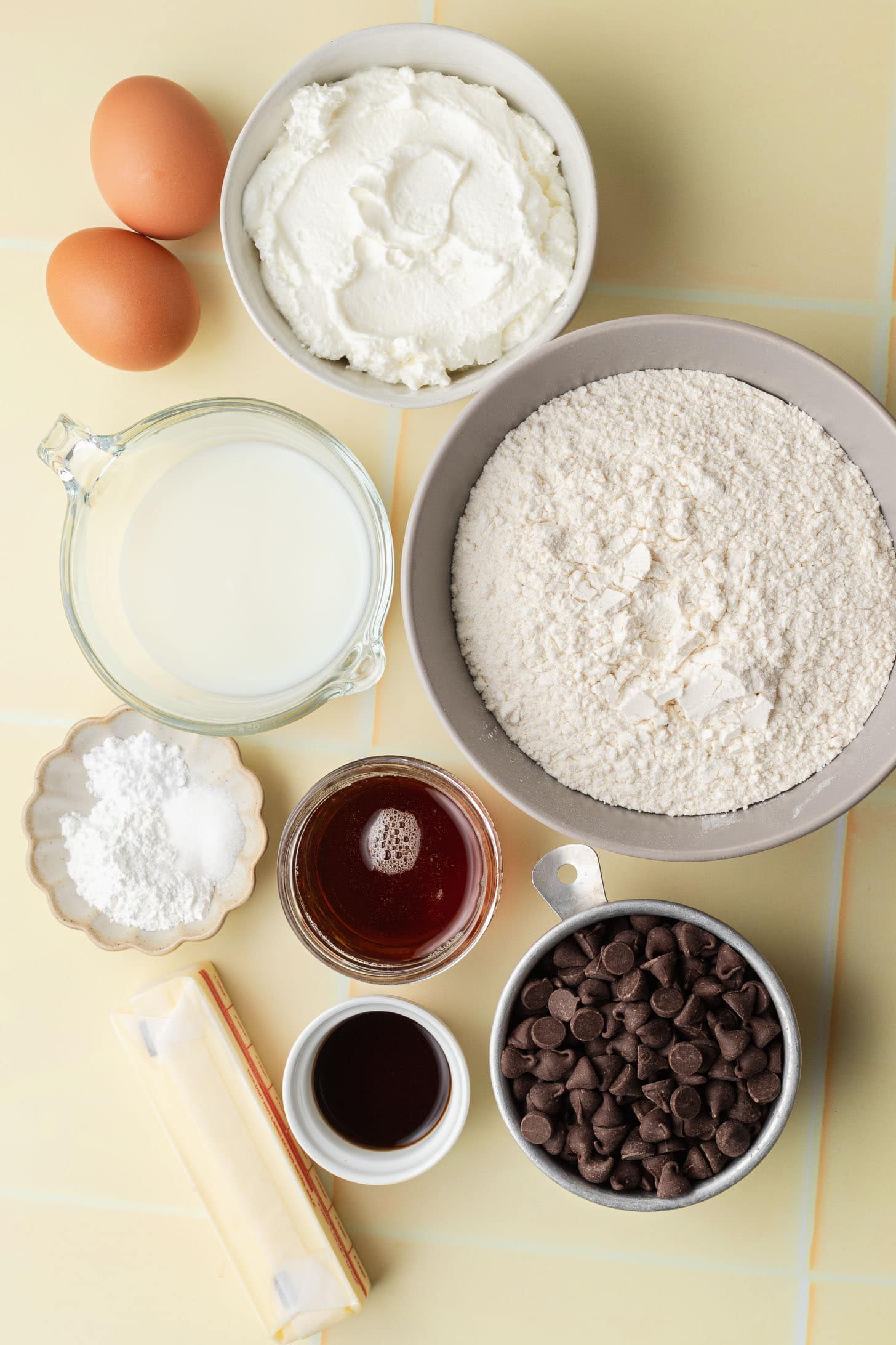 Baking ingredients in bowls: eggs, ricotta, flour, sugar, milk, butter, chocolate chips, syrup, and vanilla.