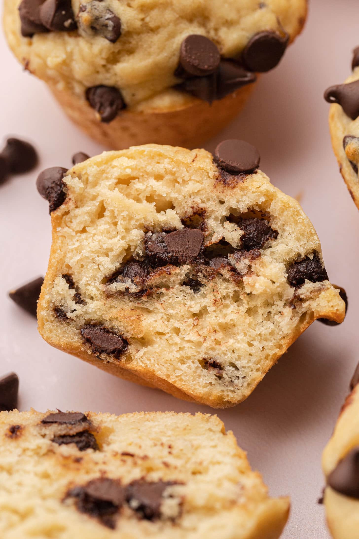 A chocolate chip muffin cut in half, showing melty chocolate chips inside, with more muffins nearby.
