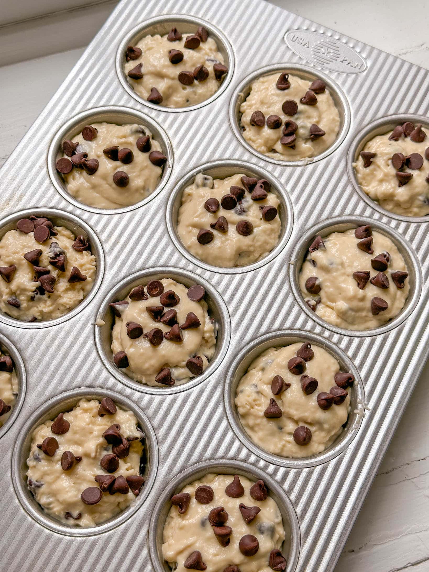 A muffin tin filled with unbaked muffin batter topped with chocolate chips, ready to go in the oven.