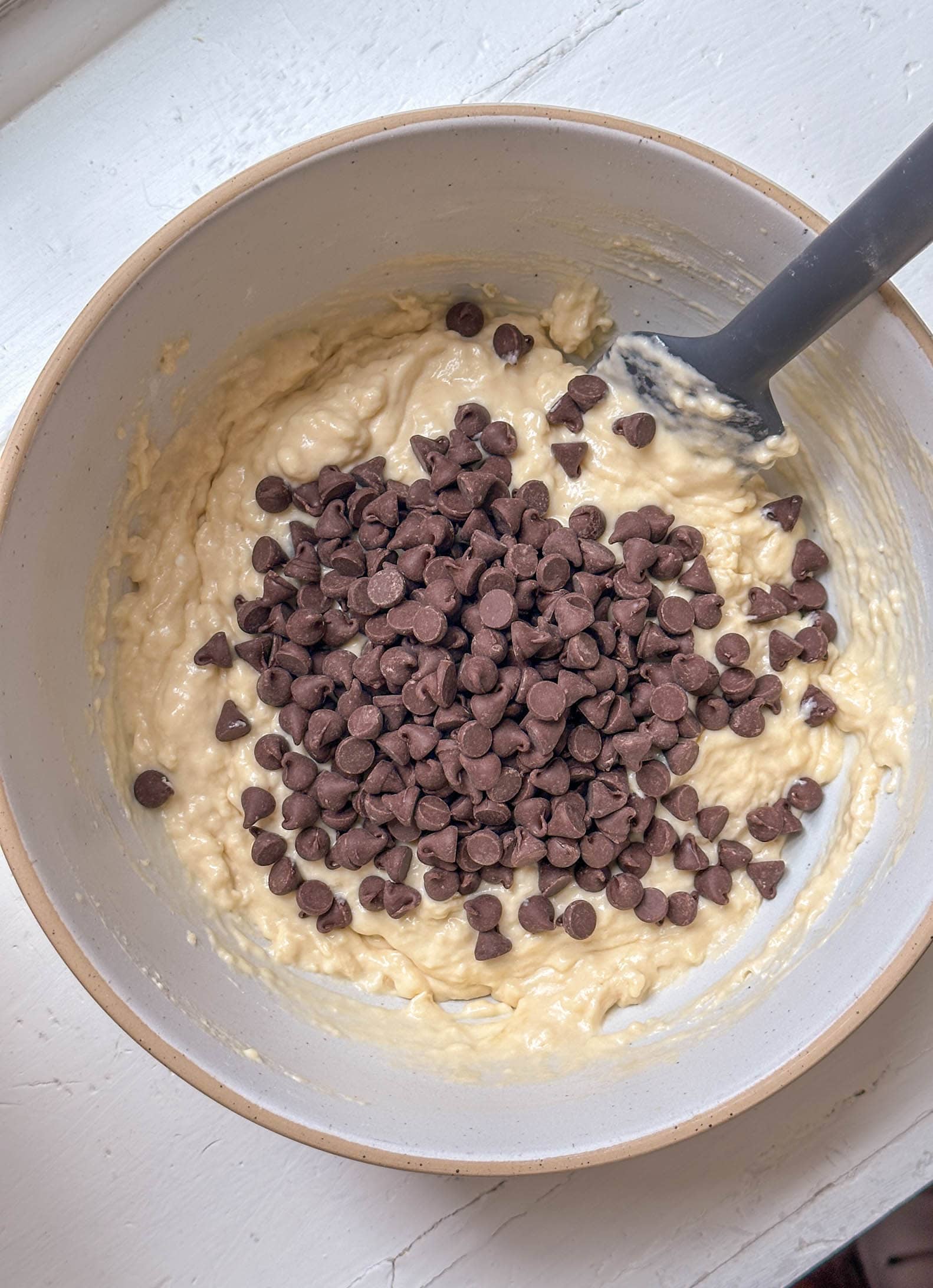 A mixing bowl with banana bread batter and chocolate chips, stirred with a spatula on a white surface.