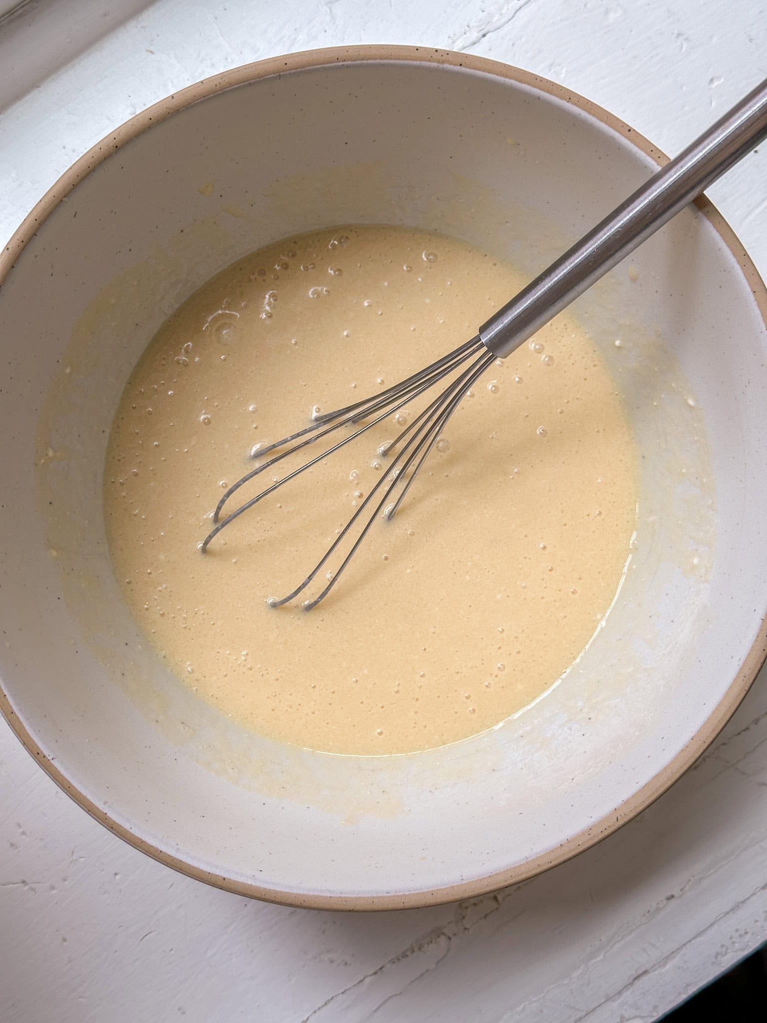 A metal whisk in a bowl of pale pancake batter on a white surface.
