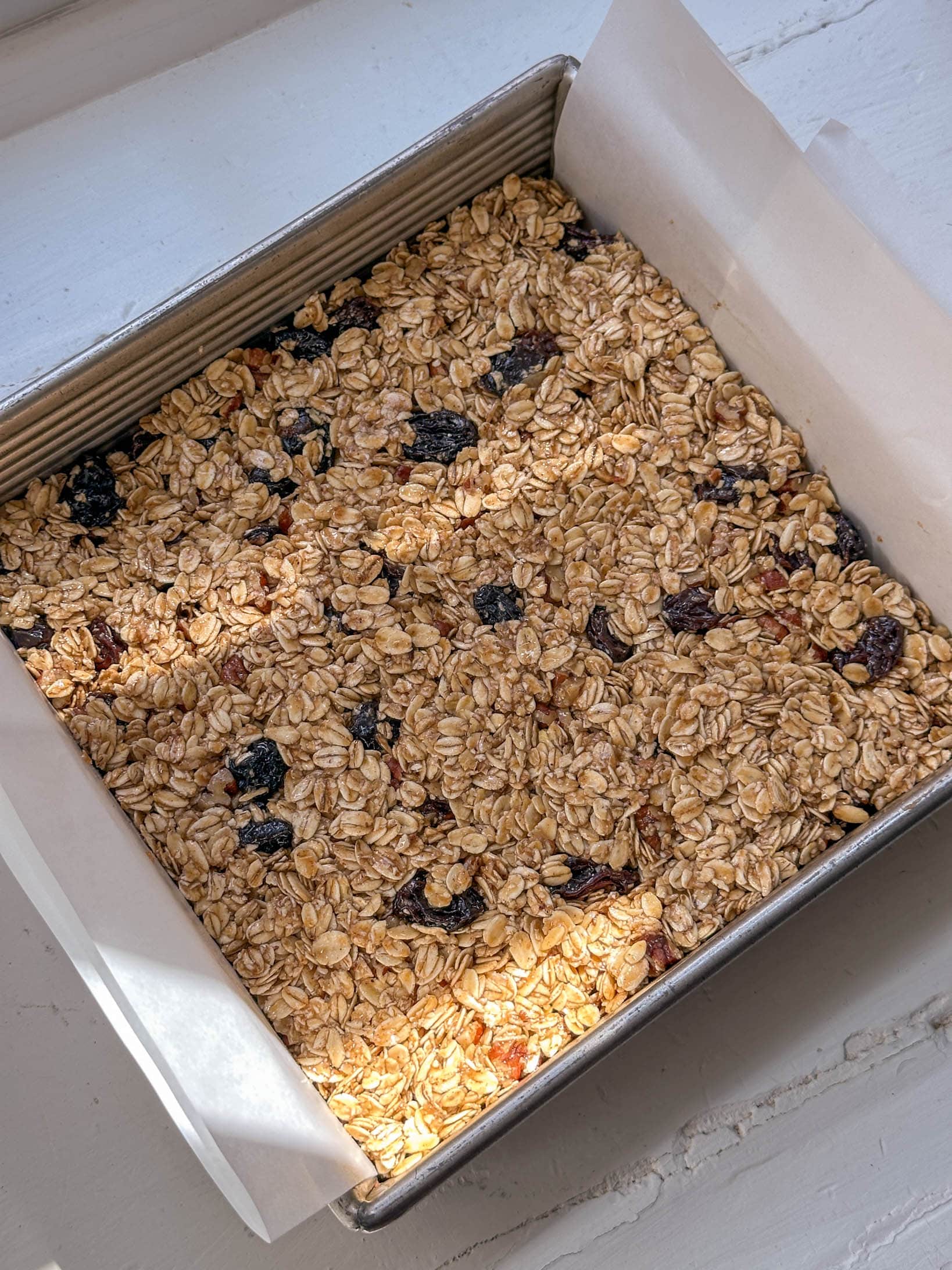 Granola bar mixture with oats and dried fruit pressed in a parchment-lined square baking pan on a white surface.
