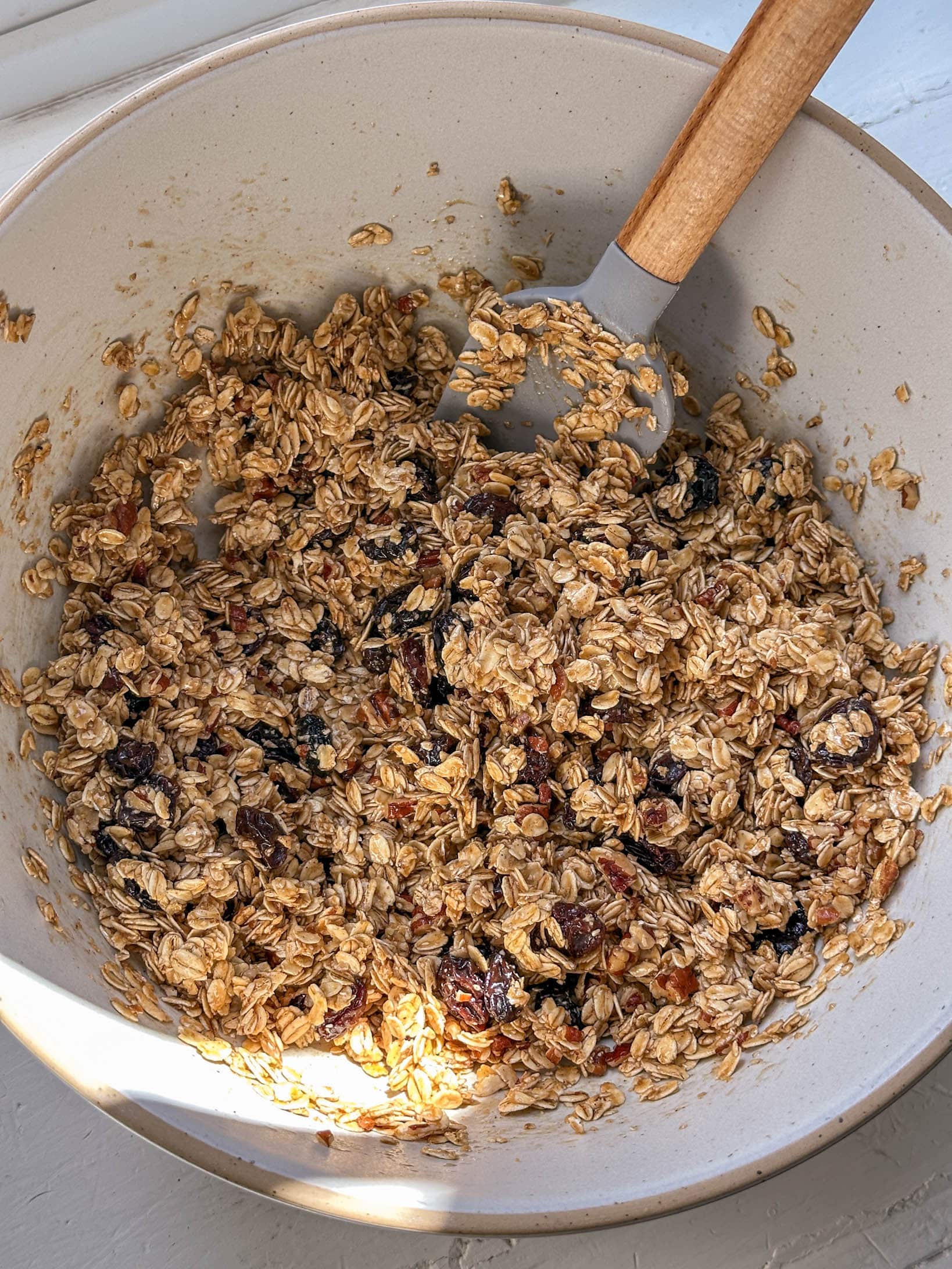 A mixing bowl with oat and dried fruit granola mixture being stirred with a spatula.