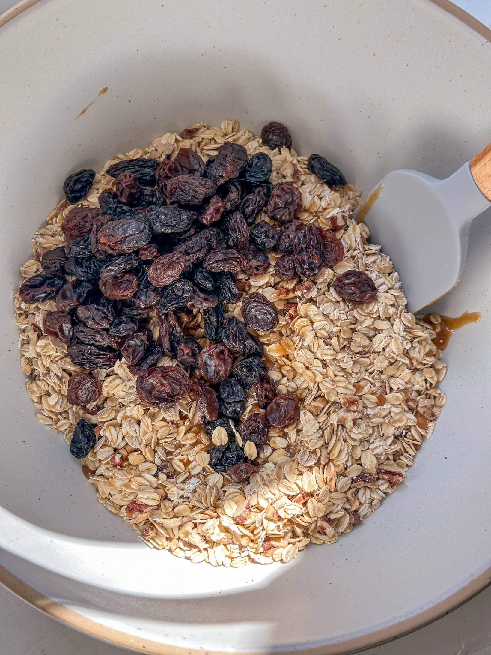 A white bowl contains oats, raisins, and seeds being mixed with a spatula on a sunny surface.
