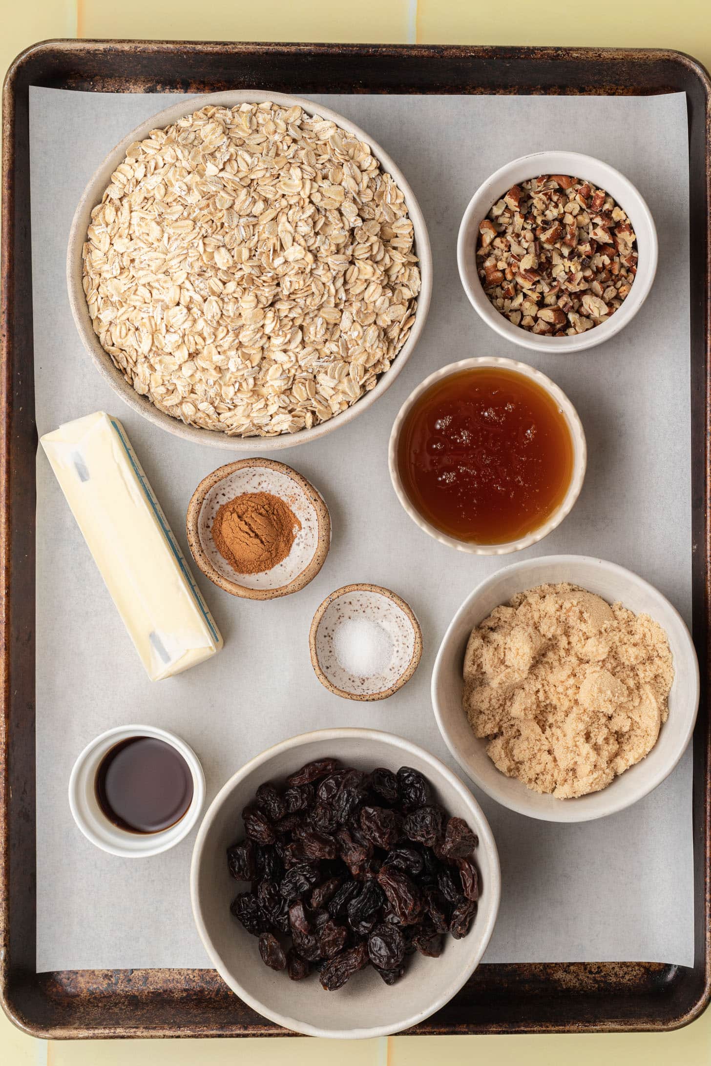 Bowls with oats, pecans, jam, brown sugar, raisins, cinnamon, salt, butter, and vanilla on a baking tray.